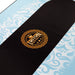 Black skateboard deck with gold logo on a blue and white patterned background