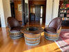 Two brown leather barrel chairs around a wooden barrel table in a room with wooden floors and bookshelves.