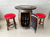 Wooden barrel table with wine storage and red cushioned stools in a room.