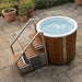 Wooden hot tub with water, ladder, and outdoor setting with potted plants.
