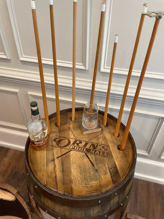 Wooden barrel with 'Drinks for a Cause' branding, surrounded by pool cues against a decorative wall.