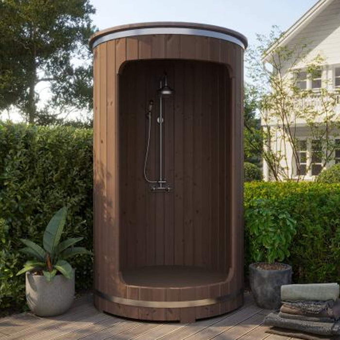 Wooden outdoor shower enclosure in a garden setting with plants and a house in the background.