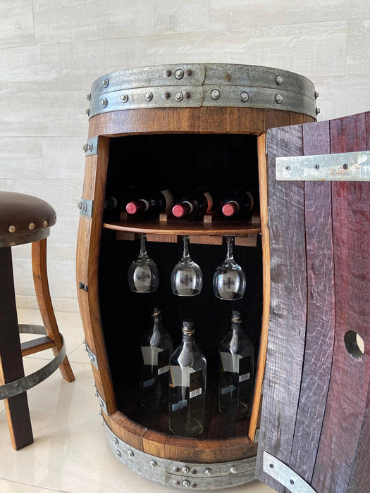 Wooden barrel with wine bottles and glasses inside, against a light-colored wall.